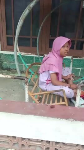 A young girl wearing a pink hijab and pajamas sits on a rusted swing set. She is in the process of playing with a red and white object, possibly a toy or candy wrapper, which she holds in her hands. The swing set is located outdoors, in front of a building with a brown window and green trim. The girl is looking towards the camera, her expression neutral. The foreground features a white ledge with a pattern of brickwork.