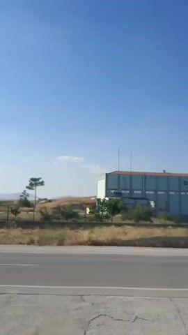 The video shows a view from a moving vehicle looking out over a highway. On the left side of the frame, a long, light blue industrial building with a white roof is visible. The building has "DSI" written in large, red letters on its side. The building appears to be raised on stilts or a platform. Beyond the building, there are green trees and a gently sloping hill. Further in the distance, the landscape is open and appears to be agricultural land with rows of crops. On the right side of the frame, a tall, lattice-like metal pylon for electricity transmission lines is prominent. The sky is clear and blue with a few wispy clouds. The road in the foreground has a gray barrier along its edge. The overall impression is of a semi-rural or industrial area viewed from a highway on a clear day.