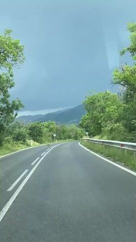 The video offers a first-person perspective from a vehicle driving along a two-lane asphalt road in Topolovo, Bulgaria. The road is marked with double solid white lines in the center and has a metal guardrail with red and white reflectors on the right. Lush green trees and vegetation densely line both sides of the road, with mountains visible in the distance. The sky is heavily overcast with dark grey clouds, giving the scene a somber or pre-storm atmosphere. Several white circular road signs with red borders are visible at intervals, providing guidance or warnings to drivers. The vehicle appears to be navigating a gentle curve in the road through this verdant, mountainous landscape.