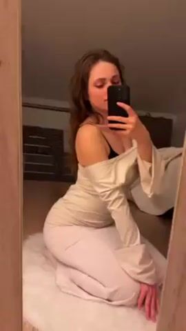A woman with long brown hair is kneeling on a white fluffy rug, taking a selfie in a mirror. She is wearing a light-colored dress with off-the-shoulder sleeves and a black bra is visible. The room has a sloped ceiling and there is a clothes rack in the background.