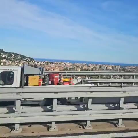 The video shows a view from a vehicle traveling on a bridge. The bridge has grey metal railings. In the distance is a city with buildings mostly of brown and white colors. Beyond the city, the blue sea meets the bright blue sky. Some wispy clouds are visible in the sky.  As the vehicle moves forward, a road sign comes into view showing a speed limit of 60 and a no-overtaking symbol.