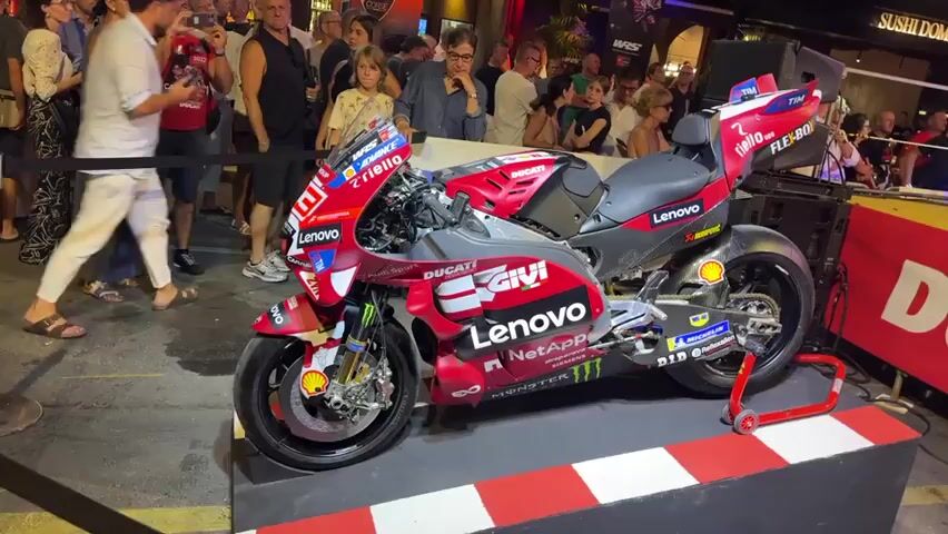 A red and gray Ducati racing motorcycle is displayed outdoors on a platform with a red and white checkered border. The motorcycle has numerous sponsor logos, including Lenovo and Givi. A crowd of people, dressed in casual attire, stands behind a black rope, watching the motorcycle. Some people are holding up their phones, possibly to take pictures or videos. The background shows a street scene with buildings and lights, suggesting it is evening or night. There is a large red banner with the Ducati logo visible to the right of the motorcycle. The scene appears to be a public event or exhibition.