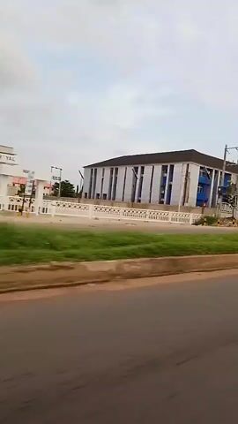 The video shows a multistory building with blue accents and white columns. A white fence runs along the front of the property, and a green car is visible driving past the building. There are utility poles and some trees in the background. The sky is overcast.
