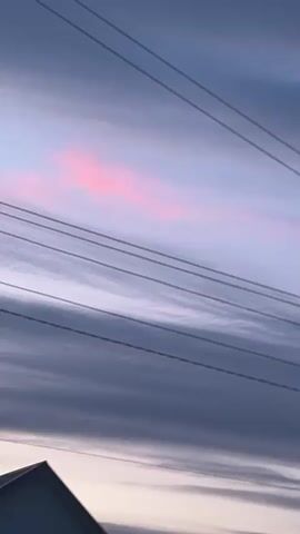 This is a vertical, close-up shot of a cloudy sky with power lines crossing the frame. The sky transitions from a light pinkish hue near the top to darker blues and grays as it moves down. Multiple power lines are visible, appearing dark against the sky and running diagonally from the top left to the bottom right of the frame. The clouds are elongated and streaky, suggesting wind. The bottom left of the frame shows a small portion of a dark, triangular object, possibly a roof or building. The overall mood is serene and somewhat atmospheric.