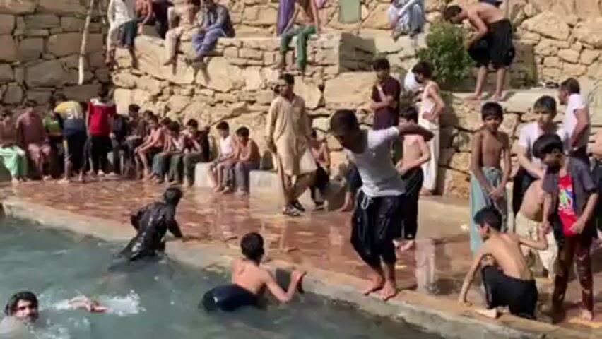 The video shows a group of men and boys swimming and playing in an outdoor pool. A large number of people are watching them from the edge of the pool and a stone wall behind it. Several people are jumping into the water, causing splashes. Some are swimming or floating in the pool. Most of the swimmers are wearing casual clothing, and some are shirtless. The pool is surrounded by tiled or stone surfaces. The environment appears to be in a rocky, rural area.