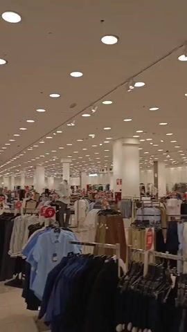 The video provides a panoramic view of the interior of a large, brightly lit clothing store, identified by "traki" signage, suggesting it is a Traki department store located in Maturin, Venezuela. The spacious store features a modern aesthetic with white walls, a white ceiling adorned with numerous recessed lights, and a highly reflective, light-colored floor.

The scene is dominated by extensive rows of clothing racks displaying a wide variety of garments, including shirts, pants, skirts, and jackets, organized into different sections. Mannequins are visible throughout, showcasing various outfits. Small red and white signs prominently display the word "traki" and the number "9," possibly indicating a price point, a section number, or a promotional offer.

The camera slowly pans across the store, revealing different clothing sections, some featuring predominantly darker colored apparel, while others showcase lighter items like blue and white shirts. The store appears relatively uncrowded, with only a few individuals partially visible in the background or behind racks, some seemingly browsing or using their phones. The overall atmosphere is clean and well-maintained, typical of a large-scale retail environment during operational hours.