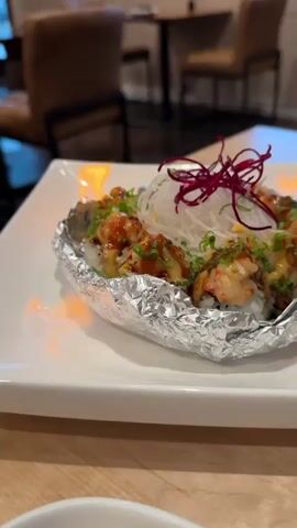 This is a close-up, vertical, eye-level shot of a flambéed sushi roll on a white square plate, which is on a wooden table. The sushi roll is served in aluminum foil, and there are flames around the sides of the foil. The sushi roll itself is topped with shredded white vegetables, red garnish, green onions, and what appears to be lobster meat drizzled with a sauce. The background is blurred but shows a restaurant setting with tables and chairs. The lighting is dim.