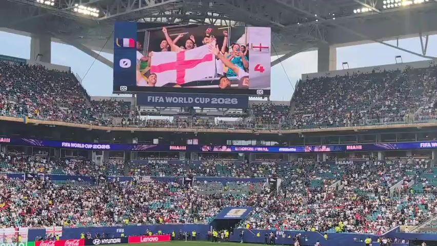 A wide-angle shot of a packed soccer stadium, likely a FIFA World Cup event, is shown. The stadium is filled with a large crowd of spectators, most of whom are wearing white and red, suggesting they are supporting England. The field is visible in the lower part of the frame, with a few players scattered across it. A large video screen at the top of the stadium displays a soccer match scoreboard showing France 0 and England 4, with images of fans holding English flags and cheering. The text "FIFA WORLD CUP 2026" is prominently displayed on the screen and also on banners around the stadium. The atmosphere appears to be energetic and celebratory.