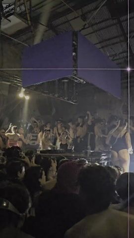 The video shows a crowded indoor dance party with a DJ performing on a raised platform. A large, cube-shaped LED screen hangs overhead, displaying abstract white and blue images. The crowd is diverse and energetic, with some people dancing and raising their arms. The DJ is surrounded by other people on the platform, some of whom are dancing as well. The venue appears to be an older industrial space with exposed beams and aged walls. The lighting is dark with spotlights illuminating the DJ and the crowd.