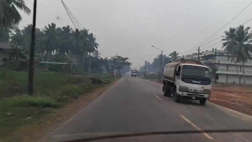 The video shows a view from a moving vehicle on a road. The road is paved and has lane markings. On the left side of the road, there is a grassy area with some vegetation and palm trees. There are also power lines running along the side of the road. On the right side of the road, there is a building with multiple floors, possibly a hotel or apartment complex. There are also palm trees and vegetation. A white truck with a dump bed is driving on the road ahead of the vehicle. The sky is overcast, suggesting it might be early morning or a cloudy day. There is a slight haze in the air. The overall scene depicts a rural or semi-urban road environment with signs of development.