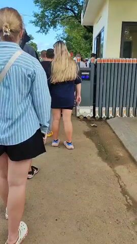 A group of people are standing in a line. The person closest to the camera is wearing a blue and white striped shirt and black shorts. The next person is a man wearing a grey shirt and black shorts. Behind him is a man wearing a black hat and black t-shirt. To the right of this man is a woman with long blonde hair wearing a dark blue shirt and black shorts. Further down the line, there are other people visible. They are standing on a paved area next to a building with a grey retractable gate. The gate has a sign that says "SAFE TRIP". There are trees and a blue sky visible in the background.