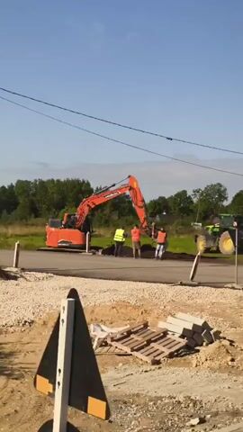 An orange excavator is being operated by a person, with four individuals standing nearby in reflective vests. They appear to be construction workers. The excavator is digging into the ground next to a paved road. There is a green tractor in the background. The environment appears to be rural with trees and fields. The sky is clear and blue. There is a pile of gravel and some wooden pallets in the foreground, suggesting ongoing construction.