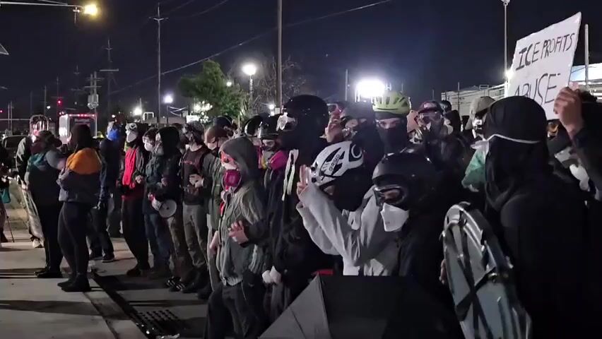 A crowd of people stand along a sidewalk at night. Most are wearing face masks or coverings, and some have on helmets. A sign reading "ICE PROFITS OFF OF ABUSE" is held by someone in the crowd. Street lights illuminate the scene.  A vehicle is visible in the background, and power lines run overhead.  The group appears to be protesting or demonstrating.