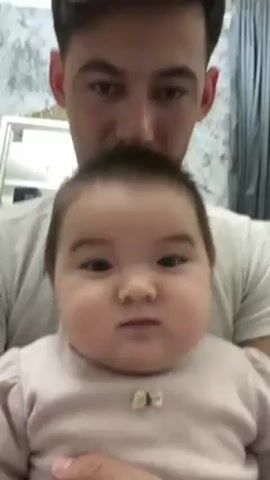 A man holds a baby, likely his child, in his arms. The baby, who appears to be less than a year old, is looking directly at the camera, making slight vocalizations. The man is looking down at the baby. The background appears to be a domestic setting with a decorated wall and curtains.
