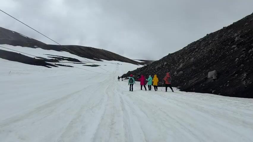 A group of hikers wearing brightly colored jackets walk along a snowy mountain path under a cloudy sky. A cable runs diagonally across the sky in the upper left of the frame. The path is covered in snow, with patches of dark volcanic rock visible on the right side of the frame. The hikers are spread out, with some closer to the camera and others further up the path. They appear to be using trekking poles.