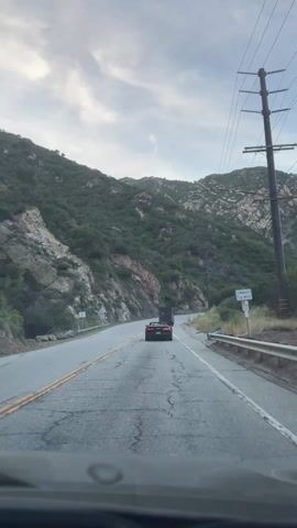 The view from inside a car follows another car on a mountain road. The road is cracked and has yellow and white lines. Mountains covered with trees are on either side of the road, and utility poles and power lines run along the right. A white car passes in the opposite direction. The sky is cloudy.