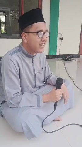 A young man wearing a black skullcap and glasses, a grey tunic, and light grey pants sits cross-legged on a white tiled floor. He holds a black microphone in his right hand and appears to be speaking into it. He is looking to his right with a slight smile. Behind him, a wall is visible with a green vertical stripe and a white section. To the left, a window with a wooden frame shows greenery outside. A black cord from the microphone trails across the floor towards the right, connected to a black electronic device with multiple cables.