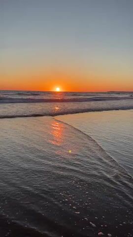 A wide shot of a beach at sunset. The sun is a bright orange orb on the horizon, casting a shimmering reflection on the wet sand. Gentle waves roll onto the shore, creating a foamy edge. The sky is a gradient of soft orange and blue.