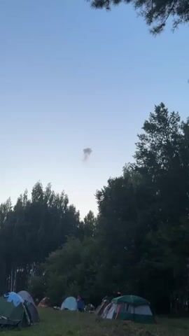 The video shows a cloudy sky with a mushroom-shaped cloud of smoke in the distance. In the foreground, there are several tents set up on grass, suggesting a campsite. Trees line the background. The audio is unclear and contains background noise, making it difficult to discern any specific dialogue or sounds.