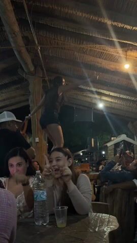 A dimly lit indoor social gathering is captured in the video. The setting features a rustic, possibly thatched roof supported by wooden beams. In the foreground, two young women sit at a wooden table. One is holding and looking at her phone with a big smile, the other is resting her chin on her folded hands, also smiling broadly. A plastic water bottle and a glass sit on the table in front of them.  In the background, several people are visible. A woman is dancing on a table, near a speaker and a small house-like structure. A man wearing a baseball cap stands next to the dancing woman, seemingly playing a musical instrument. Another man is leaning back in a relaxed pose, seemingly recording the scene with his phone. The overall atmosphere is lively and festive.
