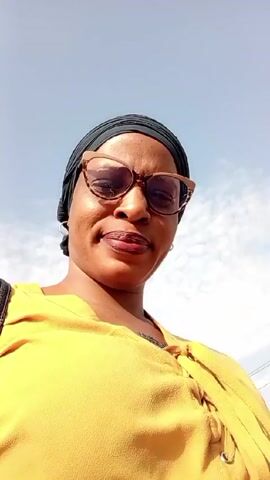 A woman wearing a yellow top, eyeglasses, and a black headscarf looks down at the camera. The background is a blue sky with scattered clouds. The video is shot from a low angle, making the woman appear taller. There are noises suggesting traffic or a busy environment.