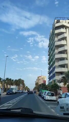 The video shows a view from a vehicle traveling down a street. The street has multiple lanes and is lined with palm trees and buildings on both sides. There are several cars and a motorcycle on the road. A large white building with balconies is visible on the right side of the street. The sky is blue with scattered white clouds. The video appears to be filmed in a city or town, possibly in a warm climate.