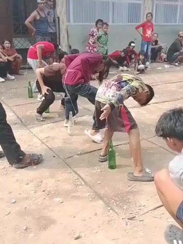 A group of children are participating in a game where they have to pick up bottles placed on the ground using their teeth. One boy is in the foreground, bent over with a bottle in front of him, and a string tied around his waist. Several other children are also bent over bottles, with strings attached to them. Spectators, including adults and other children, are watching the game from the sides. The ground appears to be paved with concrete tiles. The background shows a building with windows and doors. The lighting suggests it is daytime.