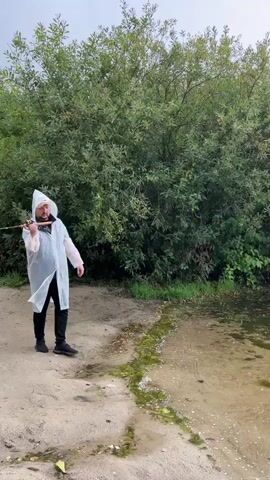 A person wearing a hooded white raincoat and black pants stands on a sandy shore next to a body of water. The person is holding a fishing rod and is in the process of casting it. Behind the person is a dense growth of green trees and bushes. The water has some green algae near the edge and the sky is overcast.