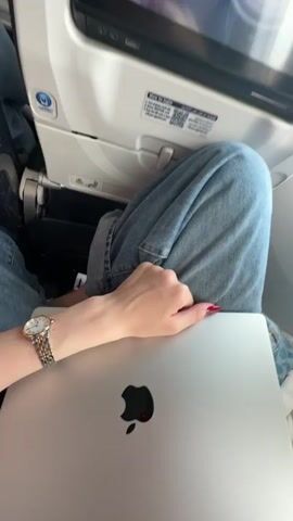 The video shows the interior of an airplane cabin. A person is seated with a laptop on their lap. The person's hands are visible, resting on the laptop. The view outside the airplane window shows clouds and a runway. The airplane is either taking off or landing. A voiceover announces that the crew is working to ensure a safe and comfortable flight.