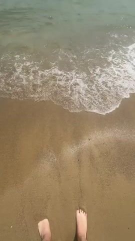 The video begins with a close-up view of a sandy beach and gentle waves washing ashore. In the bottom right corner, a bare foot is visible, indicating someone is standing at the water's edge. The camera slowly pans upwards, revealing the ocean stretching out to the horizon under a partly cloudy sky. The water is a calm green, with small waves creating white foam as they break on the sand. The overall scene is peaceful and serene, capturing a moment at the beach.