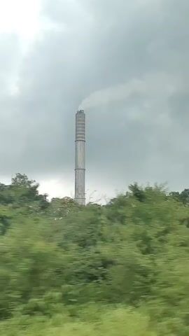 The video shows a tall, cylindrical industrial chimney emitting a plume of smoke against a cloudy sky. The chimney is situated on a rise surrounded by lush green trees. The camera perspective appears to be from a moving vehicle, as evidenced by the motion blur of the foliage in the foreground. The video captures the chimney from slightly different angles as the viewer moves past it.