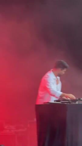 A man with dark hair and a beard, wearing a white open-collared shirt, is standing behind a DJ setup. He is looking down at the equipment, with his hands on the controls. The scene is lit with red and pink lighting, with smoke or fog in the air. The background is dark.