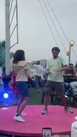 Two people, a woman and a man, are dancing on a small, round pink stage in an outdoor setting. The woman is wearing a blue dress, white sneakers, and a light-colored jacket. She has dark, curly hair. The man is wearing a light green t-shirt, dark shorts, and sneakers. He has a beard. Several other people are around the stage, watching and enjoying the performance. Some are seated, while others stand. There's a statue resembling the Statue of Liberty in the background. A camera on the ground is recording the dance. The setting appears to be on a boat or ship, with a hazy sky overhead. There is no audio.