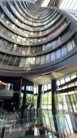 The camera pans upward from the bottom of a modern architectural structure, possibly a hotel or office building. The structure features a central atrium that spirals upward with curved balconies and multiple levels of windows. Sunlight streams in through the glass roof, illuminating the interior. In the bottom portion of the frame, there is dark-colored furniture, possibly including chairs and tables. There are several people walking in the ground floor area with large windows behind them. The exterior appears to be a park-like setting.
