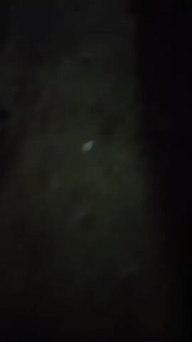 The video begins in darkness, with faint patterns suggesting nearby surfaces. A green object briefly enters the frame. A bright white light appears abruptly, obscuring the view. The light fades, and colorful lights, including blue and green, flash momentarily before the video ends. The camera appears to be in motion throughout the video.