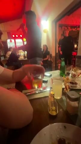 Here is a description of the video:

The video shows the interior of a restaurant or bar. A man stands in front of a group of seated patrons and sings into a microphone. He is wearing a dark jacket and light-colored jeans. Someone in the group is recording him with a phone. In the foreground, a person at the table holds a glass of amber liquid. Various other glasses, plates, and bottles are also on the table. The lighting in the room changes between red, blue, purple, and green. The other patrons are seated at tables, listening to the singer.