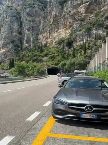 The video opens with a view of a gray Mercedes parked on the right side of a road, with its front end facing the camera. The car is in a parking spot marked by yellow lines. To the left of the car is the road, which appears to be a two-lane highway. In the background, there is a tunnel entrance in a rocky cliff face, with trees growing on the cliff. A silver SUV is parked in front of the tunnel entrance.

The video then transitions to a view from a moving vehicle. The camera is looking out the passenger side window, showing a road with a guardrail to the left. Beyond the guardrail is a body of water, likely a lake, and a steep, tree-covered mountain. The sky is blue with scattered white clouds. The camera continues to move, offering a wider view of the lake and the mountains that surround it. The sun is shining, and the water is calm. The scene appears to be a scenic drive along a coastline or lakeside.