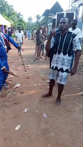 The video shows a man in a black and white patterned shirt and shorts singing into a microphone in an outdoor setting. He is barefoot and walking on a dirt ground scattered with trash. Several people are standing around him, watching and listening. Some are smiling and appear to be enjoying the performance. A large speaker is visible in the background, suggesting the event is amplified. There are houses and tents in the background, indicating a possible gathering or celebration.