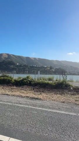 The video shows a slow pan to the right along a dry, dusty road. The road has gravel and some sparse vegetation growing on the side. In the middle ground, there is a body of water, possibly a bay or a lake, with a fence line along its edge. Beyond the water, there are some buildings and a hill covered in trees and some houses. The sky is clear and blue. The overall scene appears to be a rural or semi-rural coastal area on a sunny day.