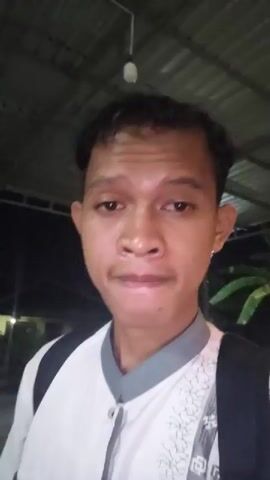 A young man with dark hair is filming himself with a phone. He is wearing a white shirt with grey trim and a backpack. He is indoors, and the background shows two people sitting on a bench and another person lying on a pillow on the floor. The lighting is dim.