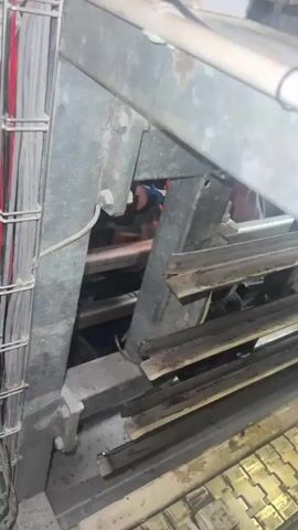The video shows a close-up view of industrial equipment, possibly within a factory or workshop setting. The primary focus is on a metal structure with a bundle of wires secured to it by a wire cage. To the right of the wires, a bright light emanates from what appears to be welding or cutting in progress. A person, wearing a white garment and possibly a cap, is partially visible on the left side of the frame, bending over and attending to the equipment. The environment seems to be dimly lit, with the bright light from the work being the most prominent light source. The overall impression is one of active industrial work being performed.