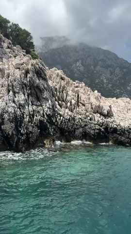 The video displays a seascape featuring a rocky coastline and clear, turquoise water. A large, predominantly white rock formation with dark accents juts out into the water on the left side of the frame. In the background, a high mountain range, partially shrouded in fog or clouds, stretches across the horizon. The sky is overcast, with a mix of gray clouds and patches of blue. The camera appears to be moving slightly, giving a sense of being on a boat or a moving platform. There is also a bird flying in the distance, away from the boat.