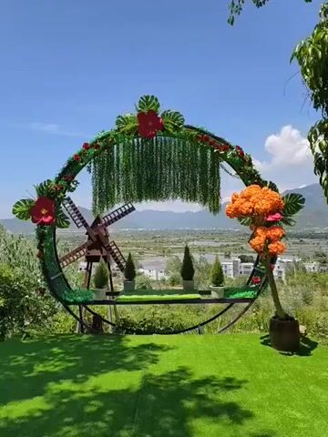 The video pans across a decorative archway set up outdoors, likely for a photo opportunity or event. The arch is circular and adorned with artificial greenery, red flowers, and cascading green vines. A small wooden windmill is positioned within the left side of the arch. Several small potted evergreen trees are placed on a shelf that spans the lower portion of the arch. To the right, a large cluster of orange artificial flowers is attached to a trunk-like base. The archway overlooks a scenic landscape with distant mountains, fields, and buildings under a clear blue sky. In the foreground, a bright green artificial turf covers the ground. There are no people visible in the video.