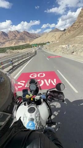 A first-person perspective video shows a person riding a motorcycle on a paved road through a mountainous, arid landscape under a clear blue sky with some clouds. The rider is wearing a black leather jacket and a helmet with a carbon fiber finish. The motorcycle is white with a camouflage pattern on the fuel tank and has several accessories attached, including a phone mount. The road has multiple lanes and a concrete barrier separating it from the mountainous terrain. In the distance, there is signage and other vehicles on the road. The mountains are rugged and rocky with sparse vegetation. The overall impression is of a scenic road trip through a remote, mountainous region.