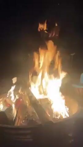 A large fire burns in a pit, the flames rising high, lit against the night sky.  Smoke swirls around the fire. A person's voice is heard on the recording.