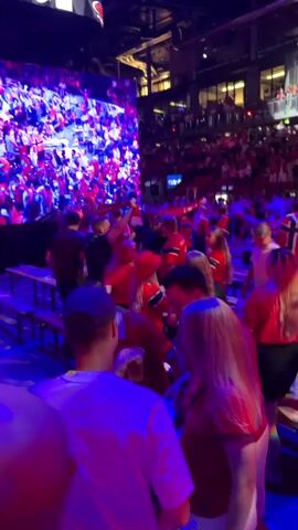 The video shows a large crowd of people gathered inside a stadium or arena. The lighting is dim and dynamic, with a combination of red and blue lights creating a vibrant atmosphere. Many of the people are wearing red clothing, suggesting they are fans of a sports team or attending an event associated with that color. Some individuals are holding up their phones, likely recording or taking pictures. The crowd appears to be standing and moving, indicating a lively event such as a concert or sporting match. In the background, elevated seating areas filled with more spectators are visible. A large screen displaying colorful lights and possibly graphics is also present, adding to the visual spectacle. The overall impression is one of an energetic and engaged audience at a public event.