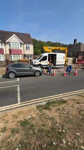 The video shows a street scene with a group of construction workers performing some kind of road work. A white van with a yellow crane is parked on the left side of the road, and a truck with a large crane is positioned in the center of the road. Several orange traffic cones are placed around the work area, and a few workers in high-visibility vests are visible. The sky is clear and blue, suggesting a sunny day. The road appears to be a residential street, with houses visible on either side. A gray car is parked on the left side of the road. There is dry grass and some weeds on the side of the road. The scene suggests that some form of utility work or road repair is taking place.