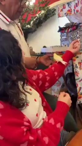 The video shows a man with glasses and a ring pointing at a small, yellow bird sitting on a woman's arm. The woman is wearing a red sweater with pink hearts and a white top with small red hearts. She is laughing. The man is standing to her left and pointing at the bird with his right hand. The background includes a decorative garland, a table covered with patterned fabrics, and other people seated in the background. The scene appears to be indoors, possibly in a cafe or shop.