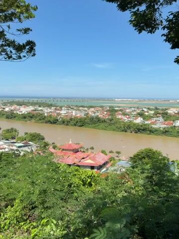 The video shows a panoramic view of a town situated along a wide, muddy river. The camera pans slowly, revealing buildings with red roofs, some clustered near the riverbank, while others spread further out. In the foreground, lush green trees and foliage frame the scene. A prominent building with a traditional architectural style, possibly a temple or pagoda with multiple red roofs and ornate details, stands near the river. In the distance, a long bridge or causeway stretches across a body of water, connecting to the horizon where the sky meets the land. The sky is a clear, bright blue. The overall impression is one of a sunny day in a riverside town.
