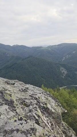 This is a vertical video showcasing a vast mountainous landscape in Sini Vrah, Bulgaria. The foreground is dominated by a large, weathered rock formation, covered in lichen, suggesting a natural, rugged environment. Beyond this, the view opens up to a sprawling valley filled with lush green forests covering rolling hills. A river or road snakes through the valley in the middle ground, with a small village or settlement visible in the distance on the slopes of the mountains. The sky is overcast with dramatic clouds, hinting at either an approaching storm or the diffused light of early morning or late afternoon. The overall mood is serene and majestic, emphasizing the grandeur of nature. There is no visible text in the video.